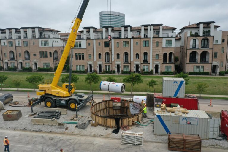 Crane lowering tunneling equipment into a circular excavation shaft at an urban microtunneling construction site.