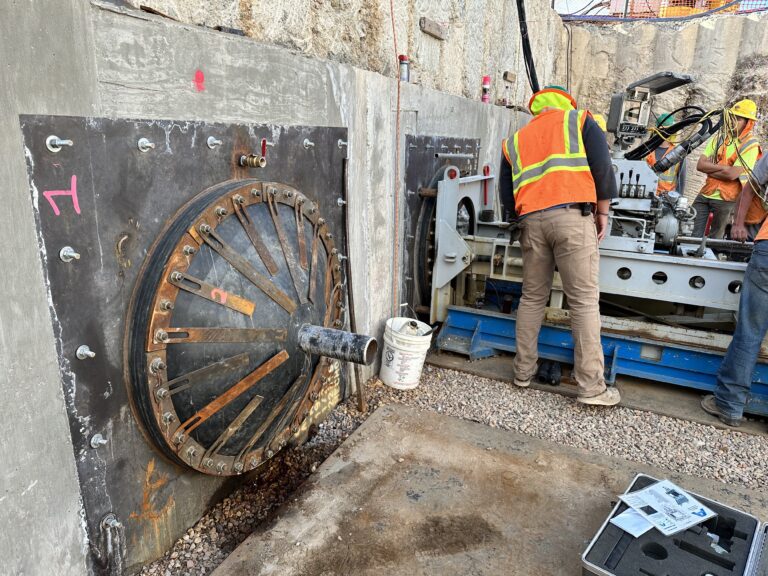 Workers operate pilot tube microtunneling equipment inside a shored excavation pit.
